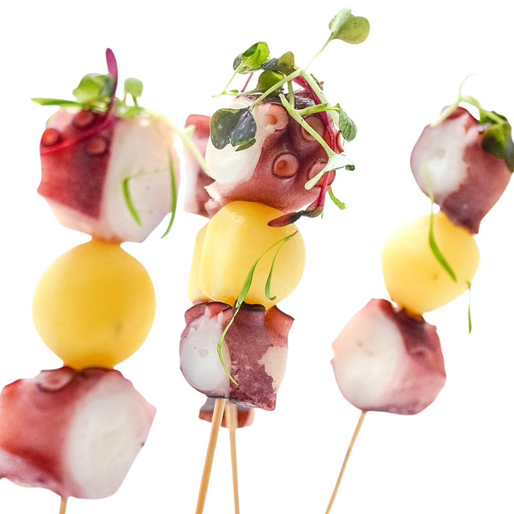 La imagen presenta unas sofisticadas brochetas de pulpo elaboradas con tiernos trozos de pulpo, patata cocida y brotes frescos como decoración. La presentación vertical y minimalista sobre un fondo blanco resalta los colores naturales de los ingredientes, transmitiendo una imagen de alta cocina y cocina mediterránea. Es una fotografía ideal para restaurantes, caterings, blogs gastronómicos, recetas de tapas y contenido relacionado con la gastronomía española y los aperitivos gourmet.