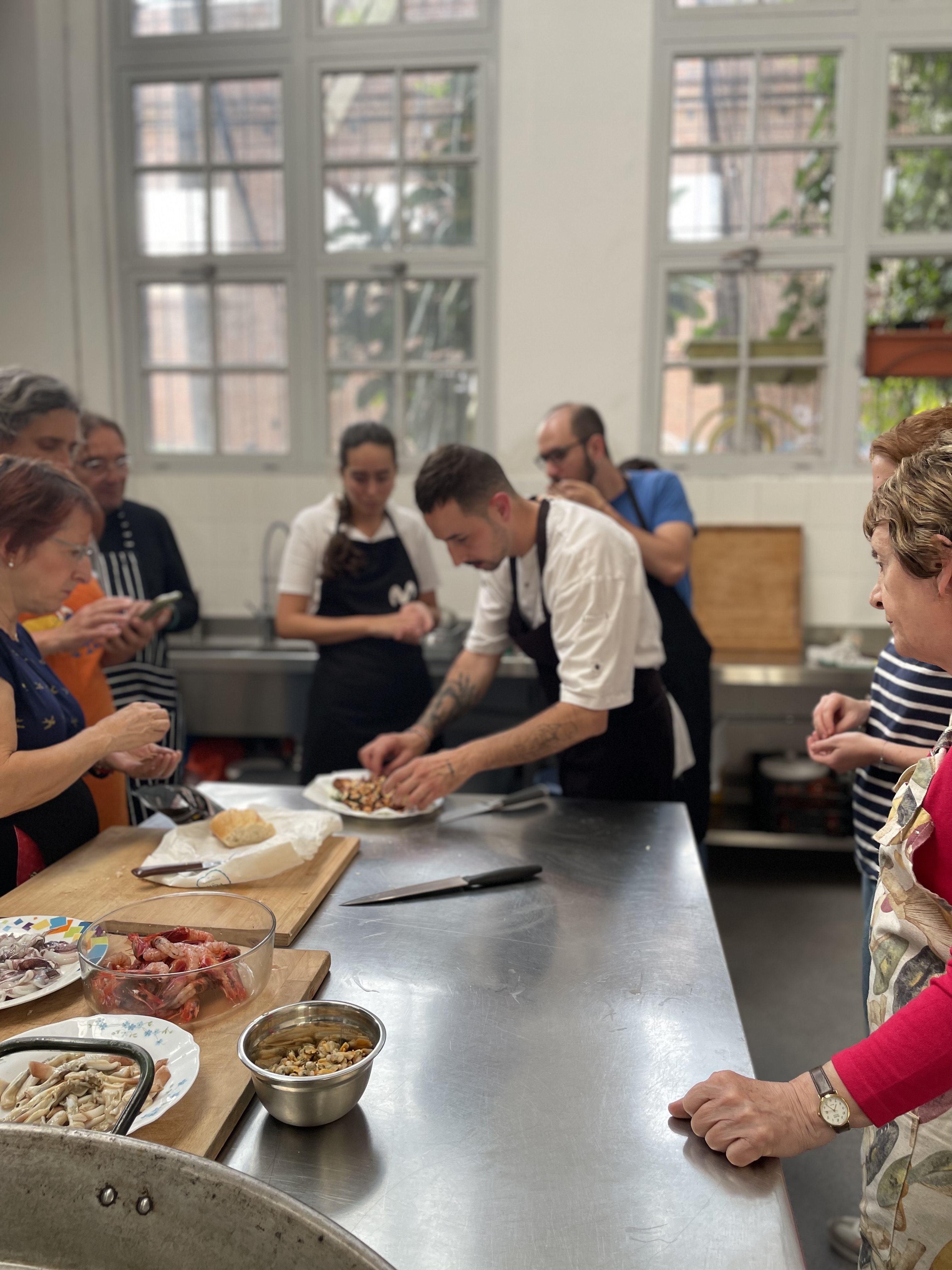 Clase de cocina del Chef Jose Cuesta en acción: un grupo de personas se reúne alrededor de una isla de cocina, con un instructor al frente dando demostraciones. Los participantes están concentrados en la tarea, que involucra varios ingredientes y utensilios de cocina esparcidos por la encimera. El entorno es una cocina interior con grandes ventanas que permiten la entrada de luz natural, creando un ambiente luminoso y acogedor para aprender habilidades culinarias.