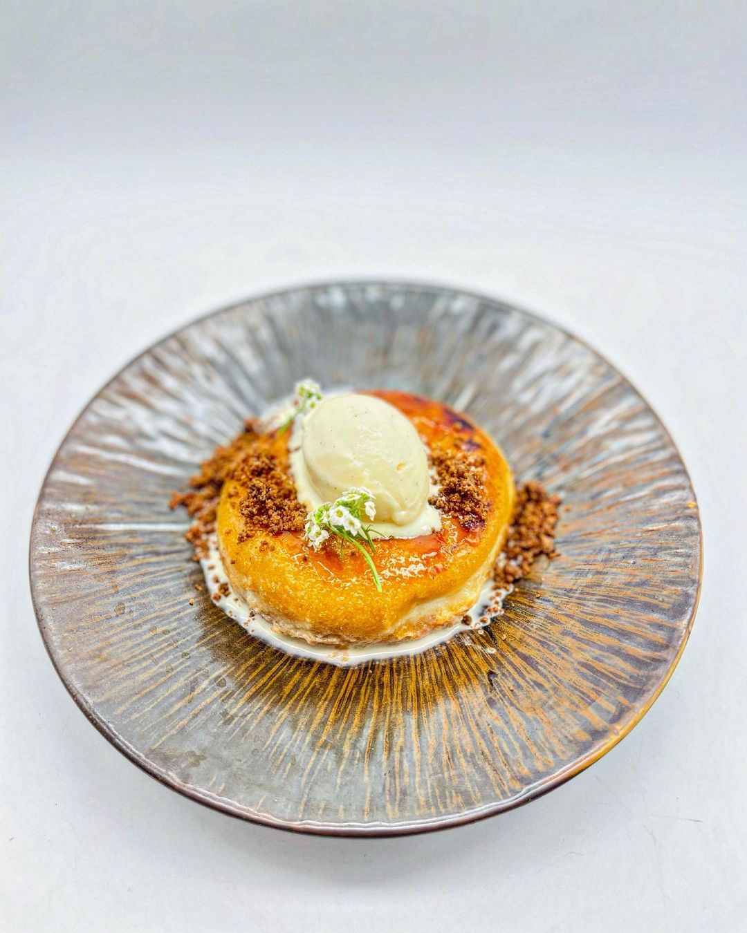 Esta deliciosa torrija de donut se encuentra en el centro de un plato de cerámica texturizada con un acabado vidriado. La parte superior de la torrija está caramelizada y presenta un color dorado. Está decorada con una bola de helado de vainilla, y se añade un toque de hierba verde para contrastar. La presentación es estéticamente atractiva, lo que la convierte en una opción visualmente interesante para entusiastas de la gastronomía o aquellos interesados en presentaciones gourmet.