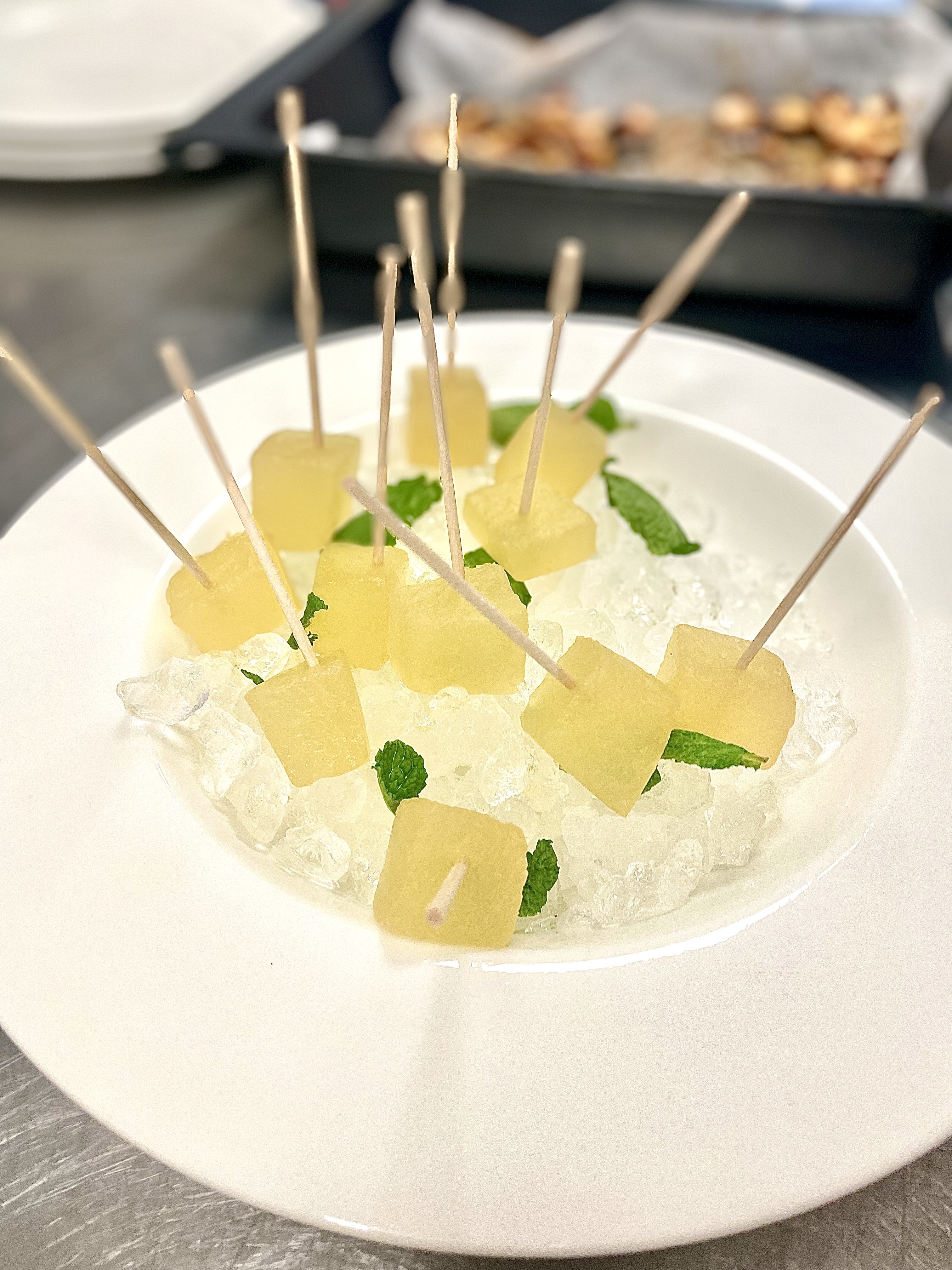 La imagen muestra una presentación culinaria de lo que parecen ser trozos de melón cocinado a baja temperatura, ensartados en palillos, adornados con hojas de menta y servidos sobre una cama de hielo picado. Esta imagen es particularmente interesante por su presentación limpia y elegante, que realza el atractivo visual del plato, lo que la hace potencialmente atractiva. La frescura sugerida por el hielo y la menta podría implica una calidad refrescante del plato, relevante para menús de verano.