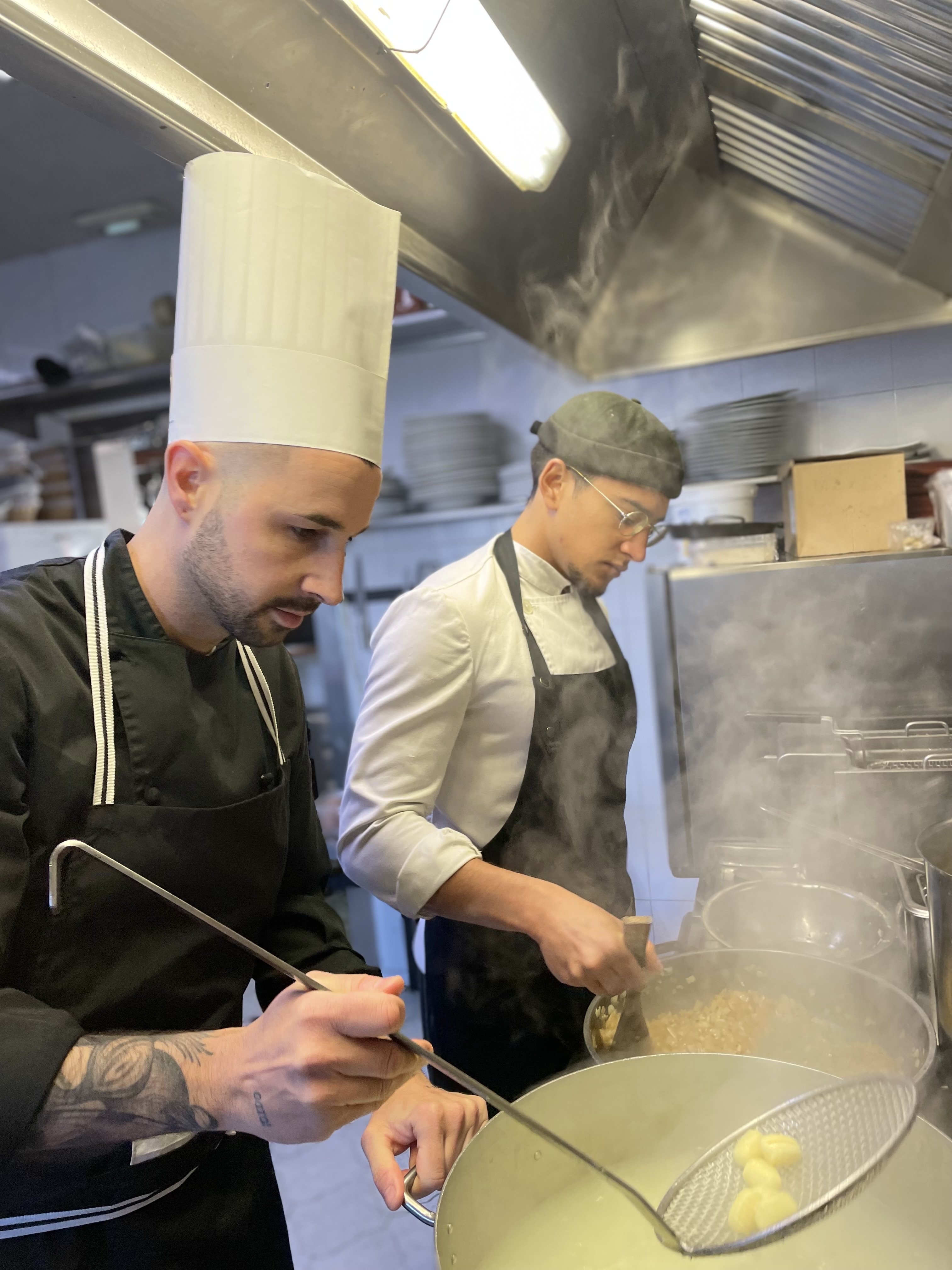 La imagen muestra a dos cocineros en un entorno de cocina profesional. La persona de la izquierda es el chef Jose Cuesta lleva un gorro y una chaqueta de chef de doble botonadura negra con botones. El individuo de la derecha lleva un delantal sobre ropa casual, lo que sugiere que es otro cocinero. El chef de la izquierda parece estar sosteniendo una sartén con comida en ella, posiblemente salteándola o sazonándola, ya que hay vapor o humo visible que se eleva de la sartén, lo que añade una acción dinámica a la escena. Esta imagen podría ser interesante o relevante para contenidos relacionados con las artes culinarias, la cocina profesional, el trabajo en equipo en la cocina.