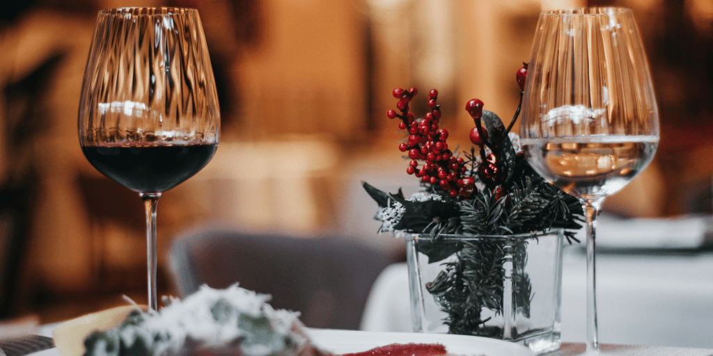 Mesa navideña en un ambiente cálido, vestida con mantel blanco hay dos copas de vino tinto, tras ellas un adorno elegante de acebo navideño.