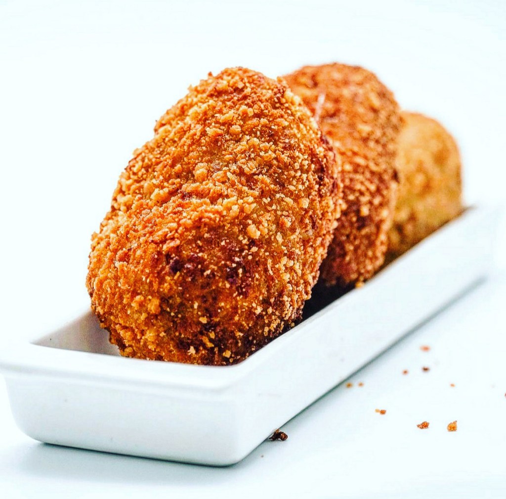 La imagen muestra una presentación de croquetas empanizadas y fritas, servidas en un plato rectangular blanco. Las croquetas tienen una apariencia dorada y crujiente, lo que sugiere que han sido bien cocinadas y tienen una textura apetecible. Están dispuestas en fila, resaltando la atención en la presentación. Este tipo de platillo es típico en la cocina de aperitivos y puede contener diversos rellenos, como jamón, pollo o queso.