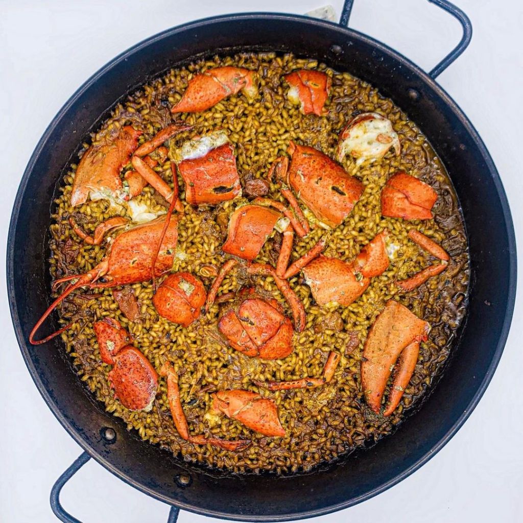 La imagen muestra una auténtica paella de mariscos con trozos de bogavante sobre arroz meloso, cocinada a la perfección en paellera tradicional. Un plato mediterráneo ideal para compartir en reuniones familiares o eventos especiales, que destaca por su sabor y frescura. Perfecto para los amantes de la cocina española y el marisco.
