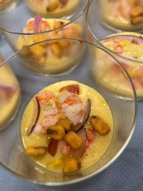 La imagen muestra un delicioso cóctel de mariscos en vaso individual, con camarones frescos, cebolla morada en finas tiras y crocantes trozos de crujiente maíz tostado, servido sobre una cremosa base de salsa. Perfecto como aperitivo en eventos o reuniones. Ideal para los amantes de la cocina gourmet y los sabores del mar.